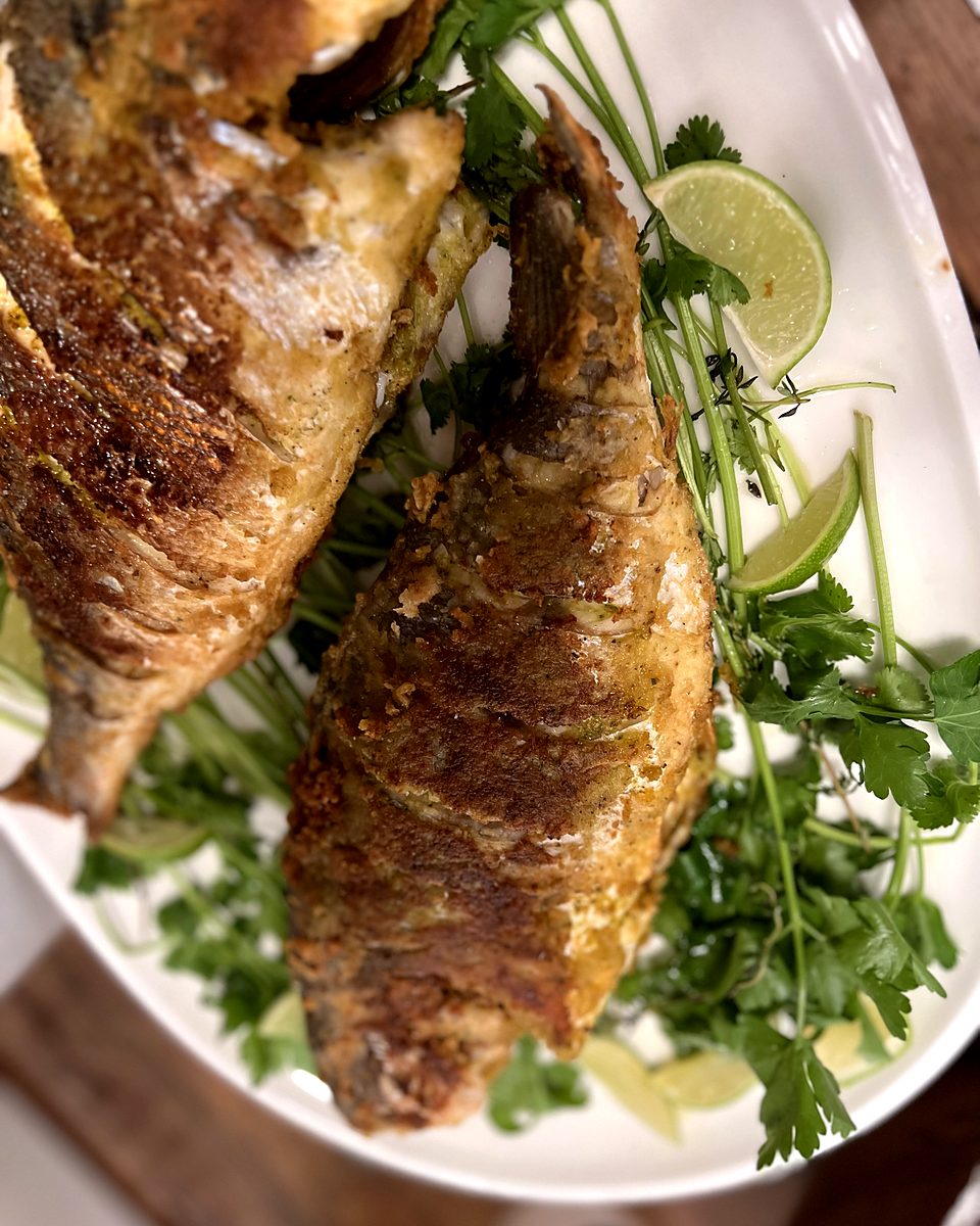Whole fried fish on a white platter with lime wedges and fresh herbs